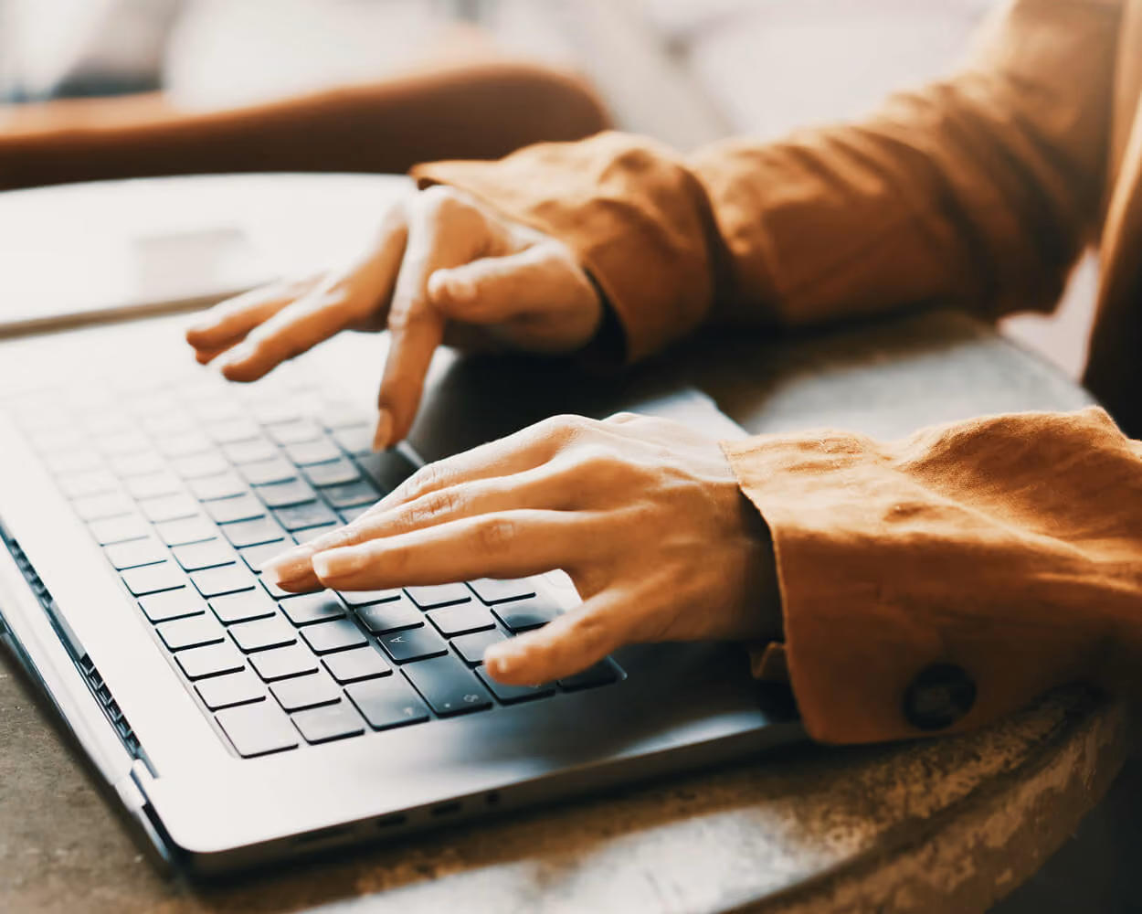 Close-up of a person's hands typing on a laptop keyboard, wearing a mustard-colored shirt, resting on a concrete surface.