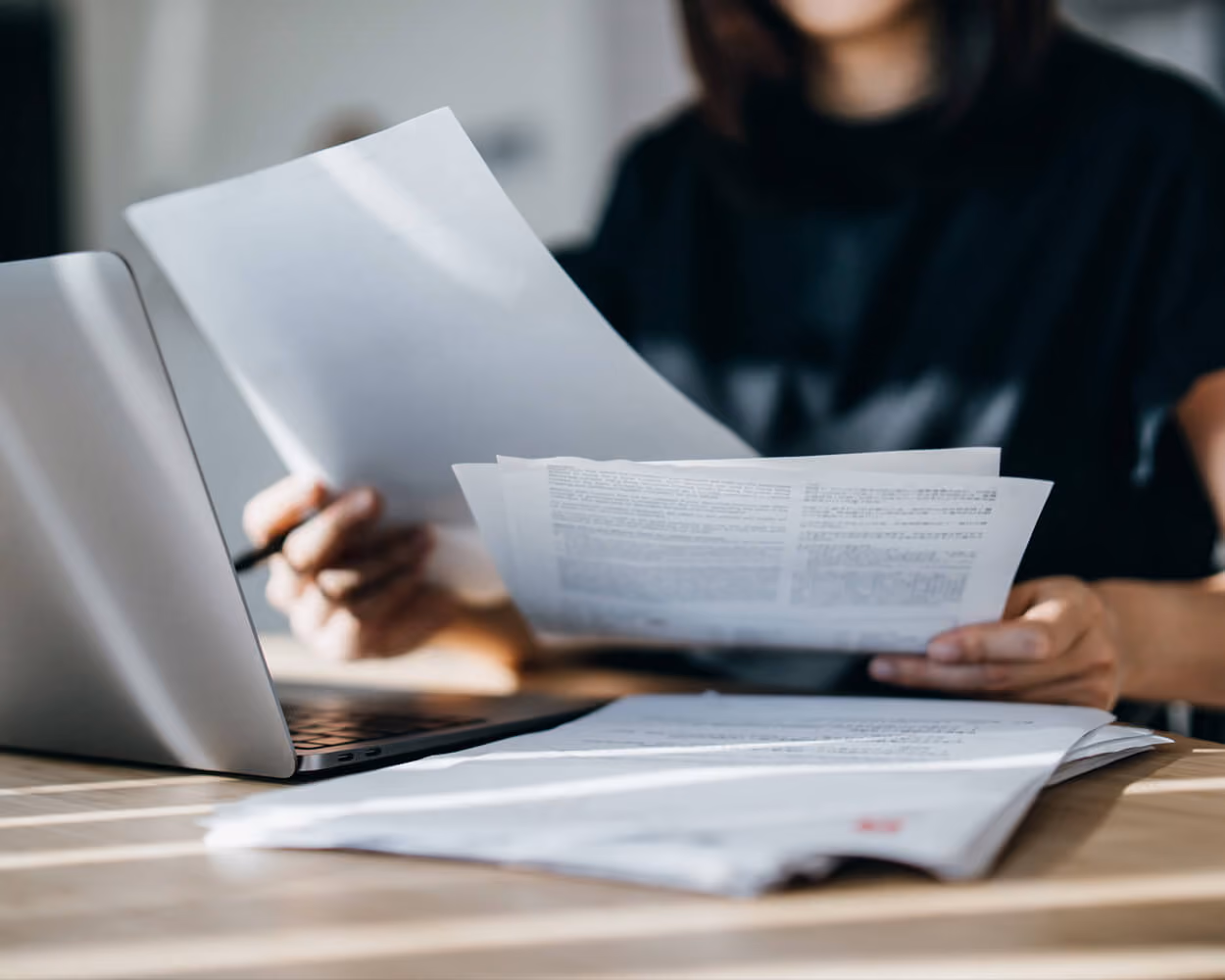 Person seated at a table holding a printed document in one hand and a pen in the other, reading it beside an open laptop, with a stack of papers on the desk.