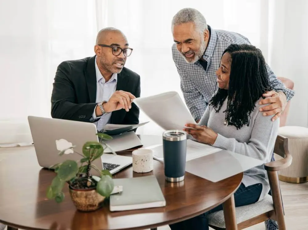 Lawyer explaining estate planning documents to a mature married couple.|Estate planning documents with reading glasses laying on documents.|Woman smiling as she holds couple's estate planning documents while standing next to seated husband and lawyer.|Estate planning documents and a pair of reading glasses.|Woman smiling as she holds couple's estate planning documents while standing next to seated husband and lawyer.