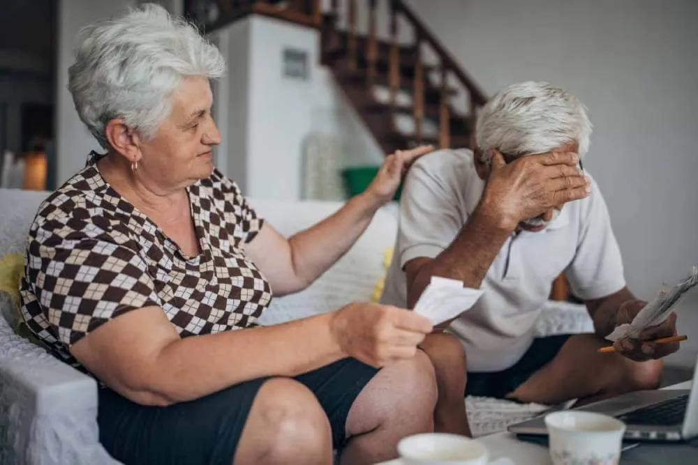 Elderly wife comforting husband after experiencing an elder financial exploitation event.|Elderly husband and wife reviewing receipts after being financially exploited.|Elderly husband and wife worried after experiencing elderly financial exploitation.|Elderly couple reviewing receipts after experiencing elder abuse.|Worried couple concerned about elder abuse.