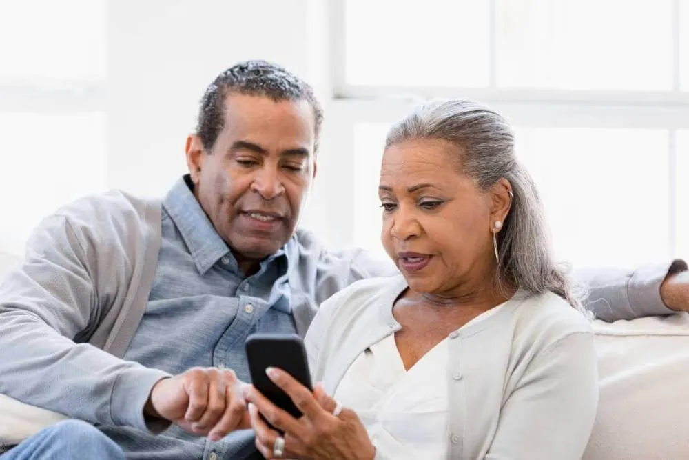 Couple looking at phone as they discuss retirement.|Husband pointing at retirement planning documents as wife looks on.|Couple looking at retirement documents while sitting in front of a laptop computer.|Retirement planning couple reviewing legal documents.|Couple reviewing retirement planning documents.|Couple looking at retirement planning documents and their laptop computer.|Retirement planning document reivew by mature couple.