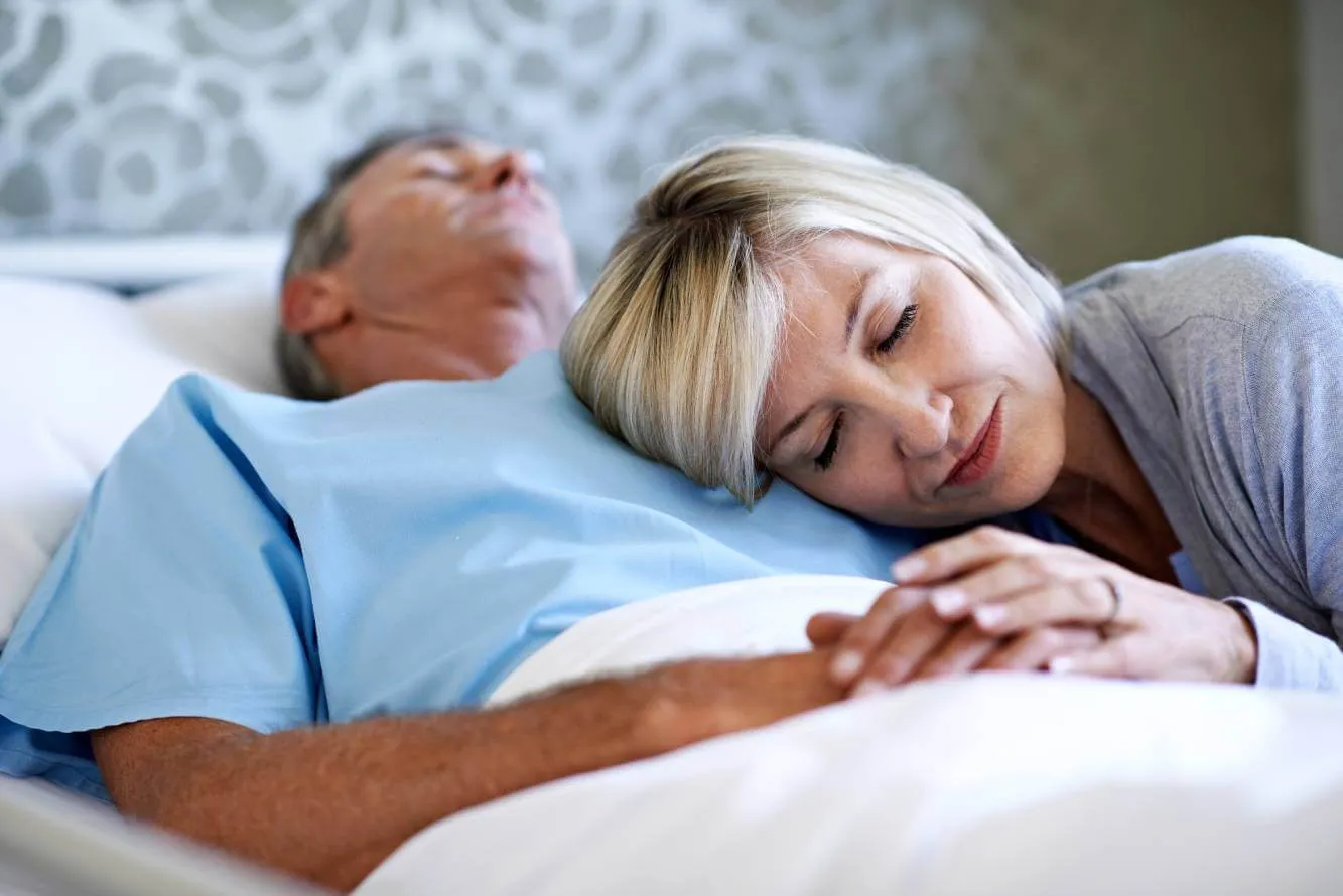 Wife laying her head on husband who is incapacitate in a hospital bed