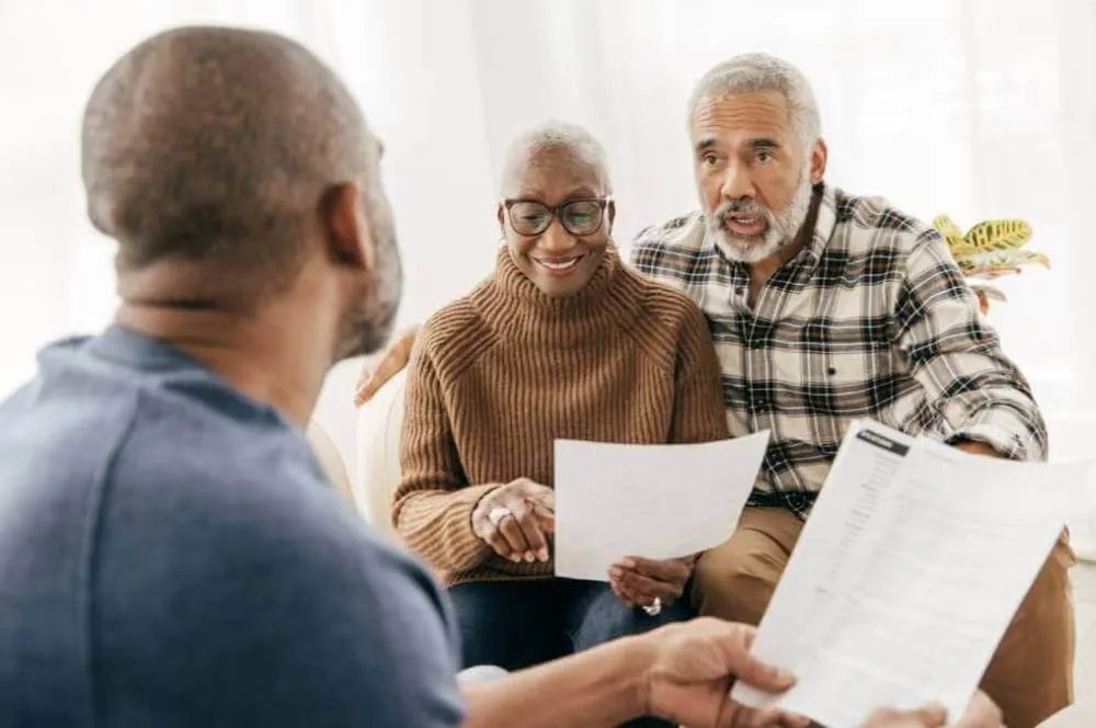 An older couple getting a legal consultation from a younger man.|A man explained an Elder Law issue to an older couple while standing in a living room.|A lawyer explaining an legal elder law issue to an elderly couple in their living room.|Lawyer answering questions about elder law for an elderly couple.|Son explaining elder law to elderly parents.