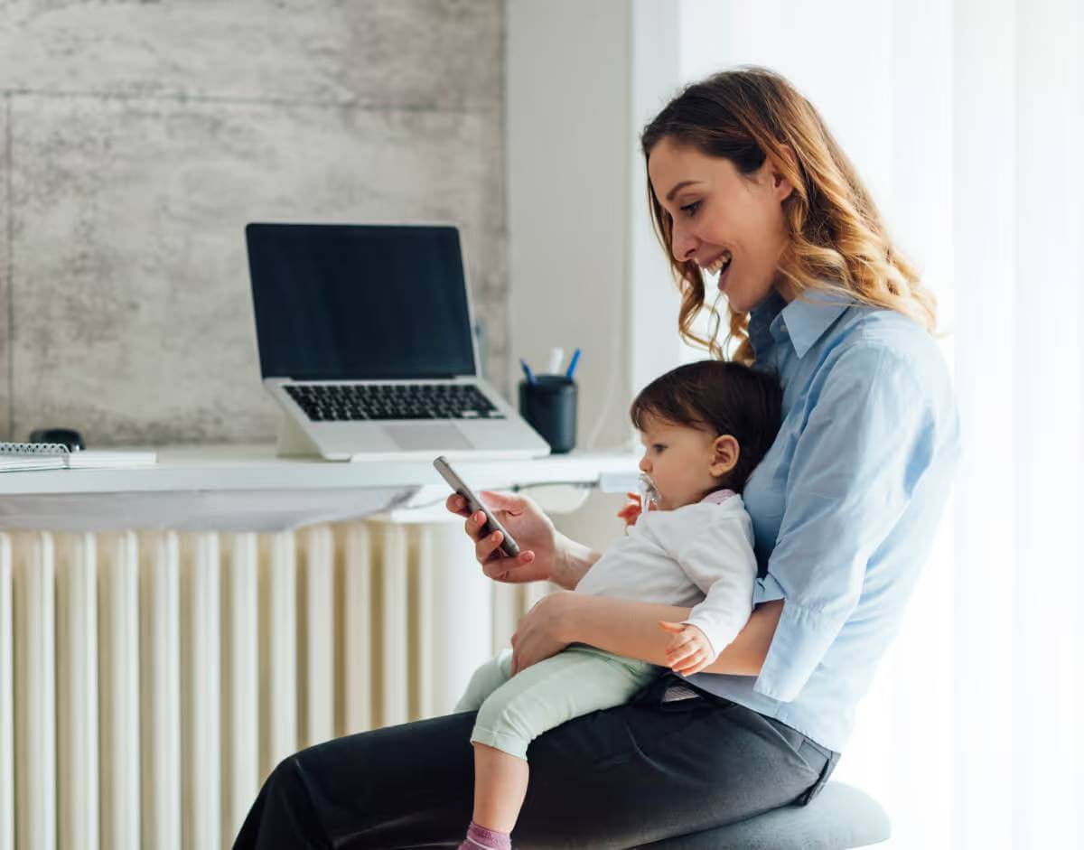 Mother holding her child as she looks at mobile phone and smiles.