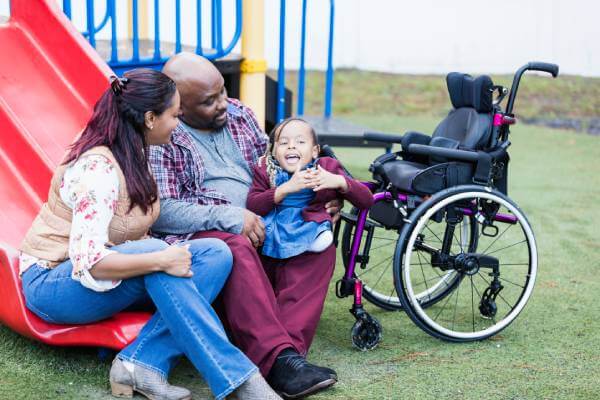 parents talking their young special needs child while sitting on a playground slide