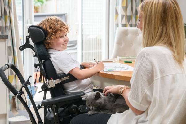 Young boy ins an electric wheelchair doing school work at a dining room table as he smiles at his mother