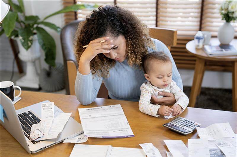A woman with head down who appears stressed by bills, paperwork that surrounds her. She is holding a baby.