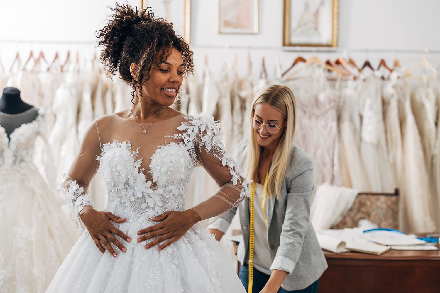 A bride trying on a wedding dress in a shop, with the vendor measuring behind.
