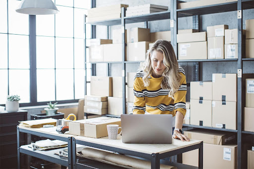 A person works on a laptop in a stock room with boxes lining the shelves, choosing the right type of business address for LLC.