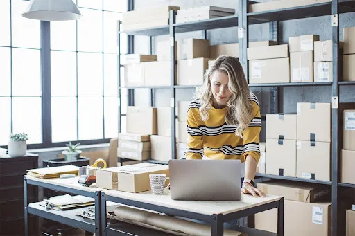 A person works on a laptop in a stock room with boxes lining the shelves, choosing the right type of business address for LLC.
