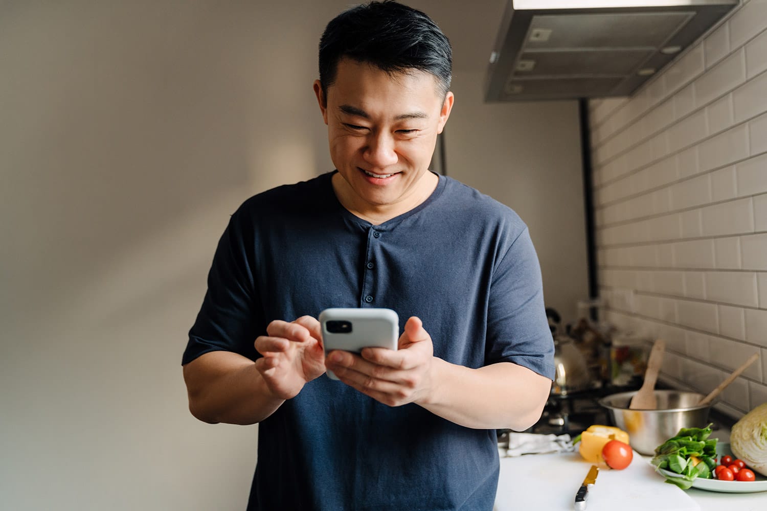 A man standing in his kitchen, leaning on a table, looking at his laptop and a document.