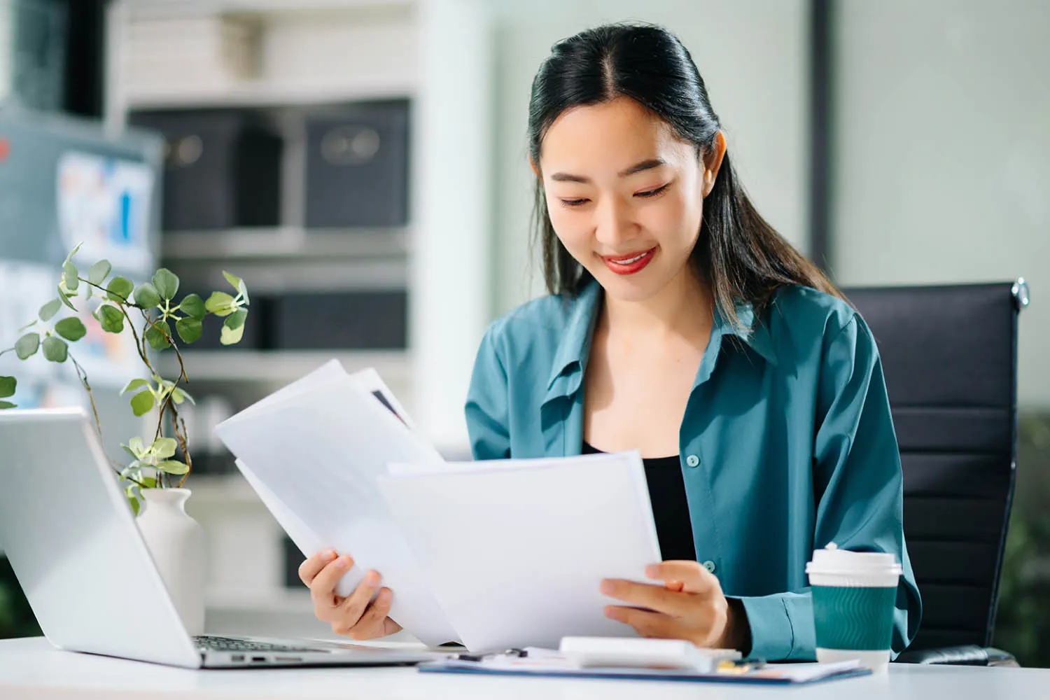 A professional businessperson sits at a desk using a laptop or reviewing paperwork, smiling in an office environment.