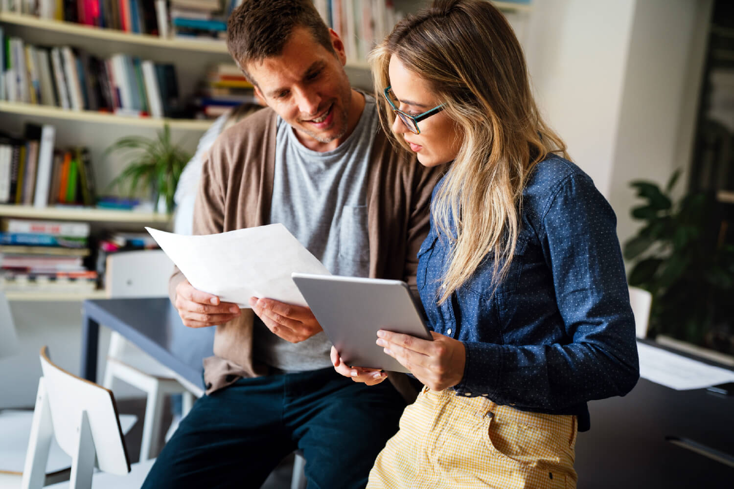 Two people convene to review a sheet of paper and a tablet.
