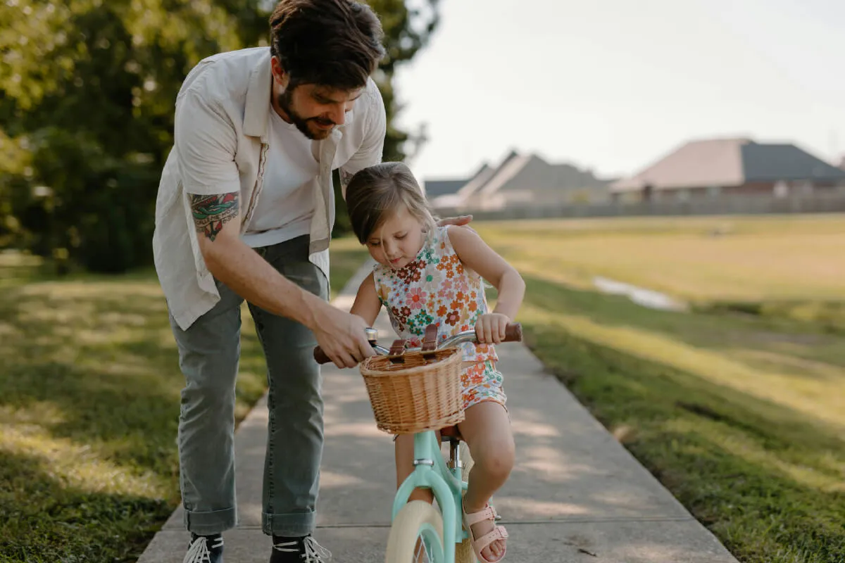 A parent helps a small child ride a bicycle on a shady sidewalk.