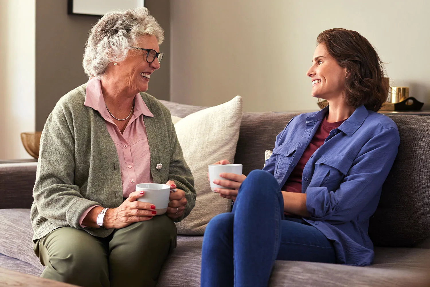 A woman and her mother having tea together and smiling at one another.