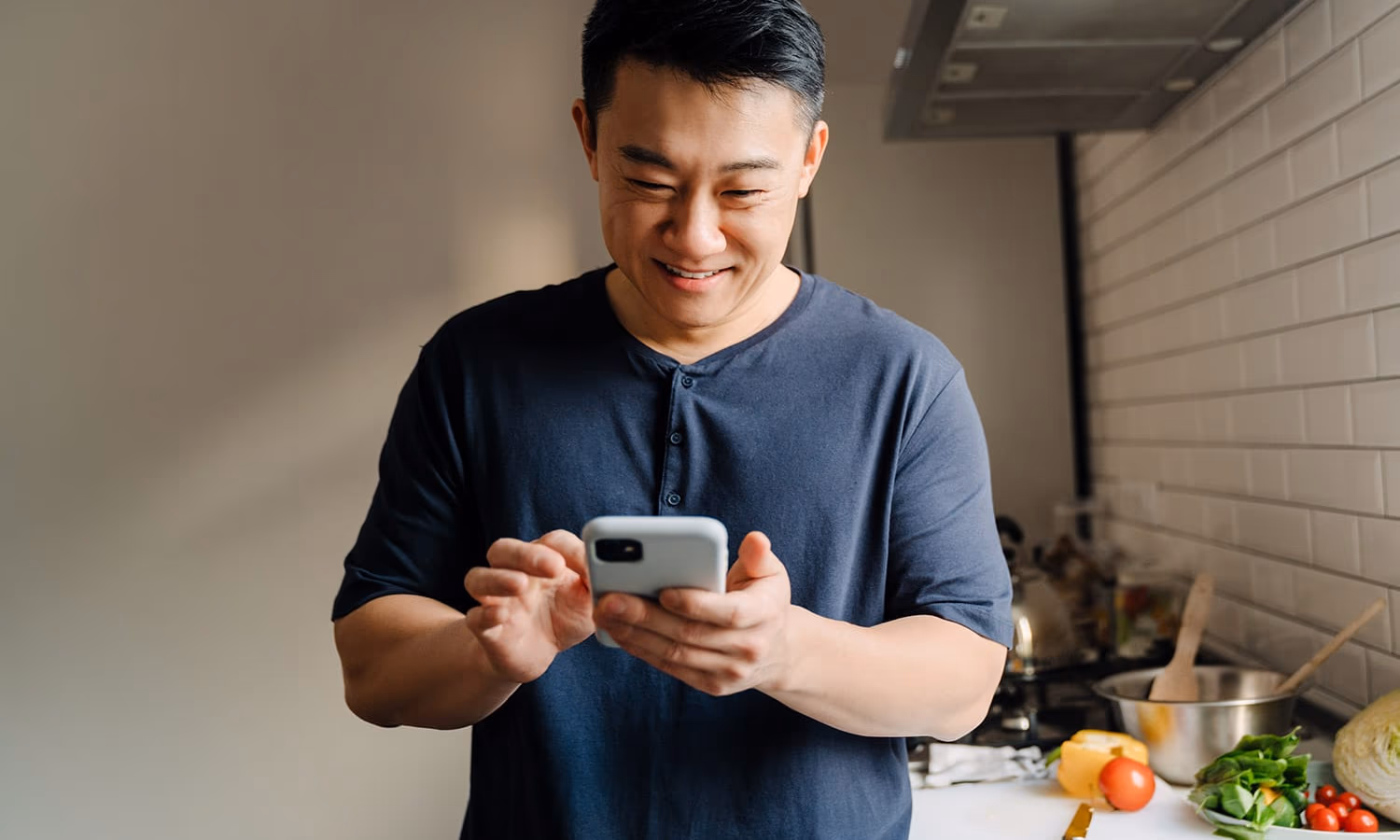 A man standing in his kitchen, leaning on a table, looking at his laptop and a document.