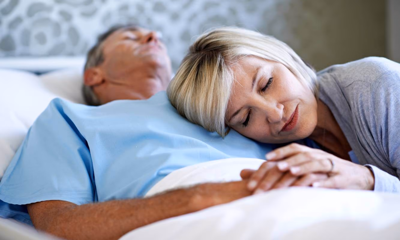 Wife laying her head on husband who is incapacitate in a hospital bed