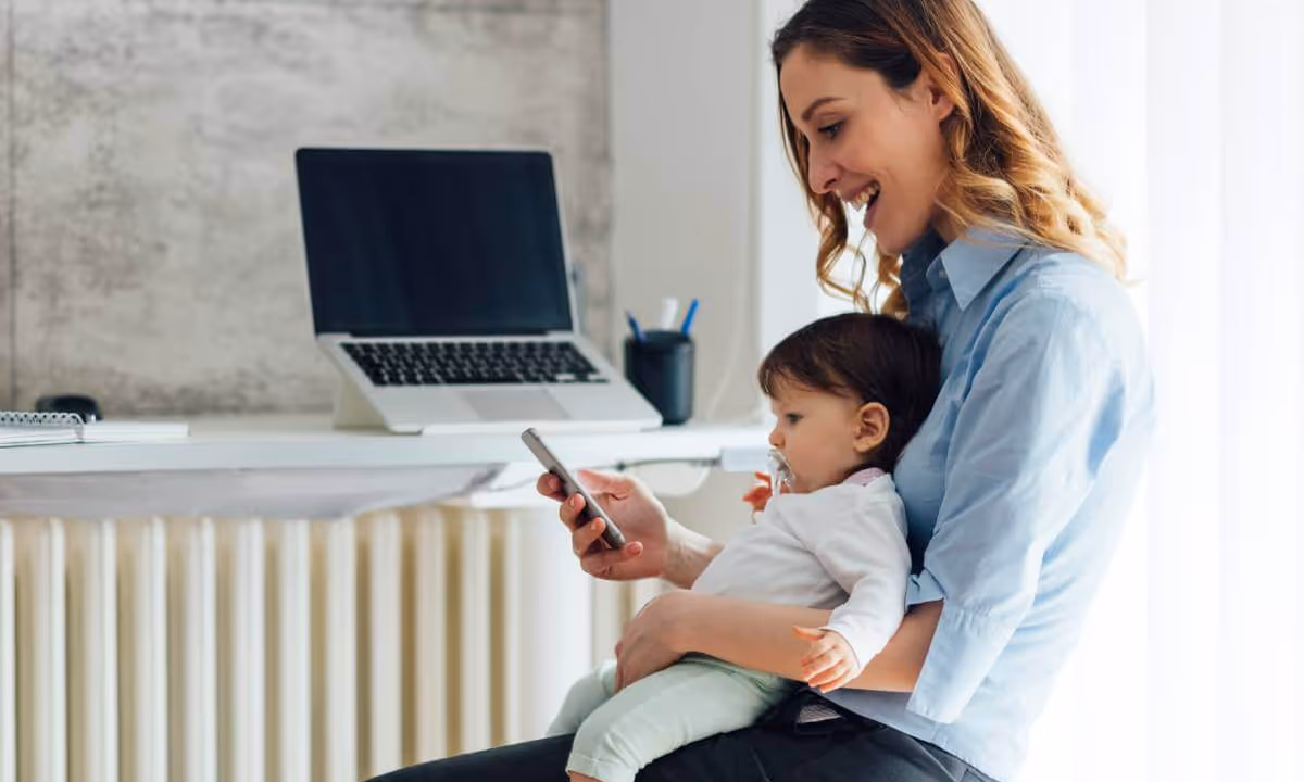 Mother holding her child as she looks at mobile phone and smiles.