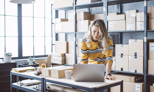 A person works on a laptop in a stock room with boxes lining the shelves, choosing the right type of business address for LLC.