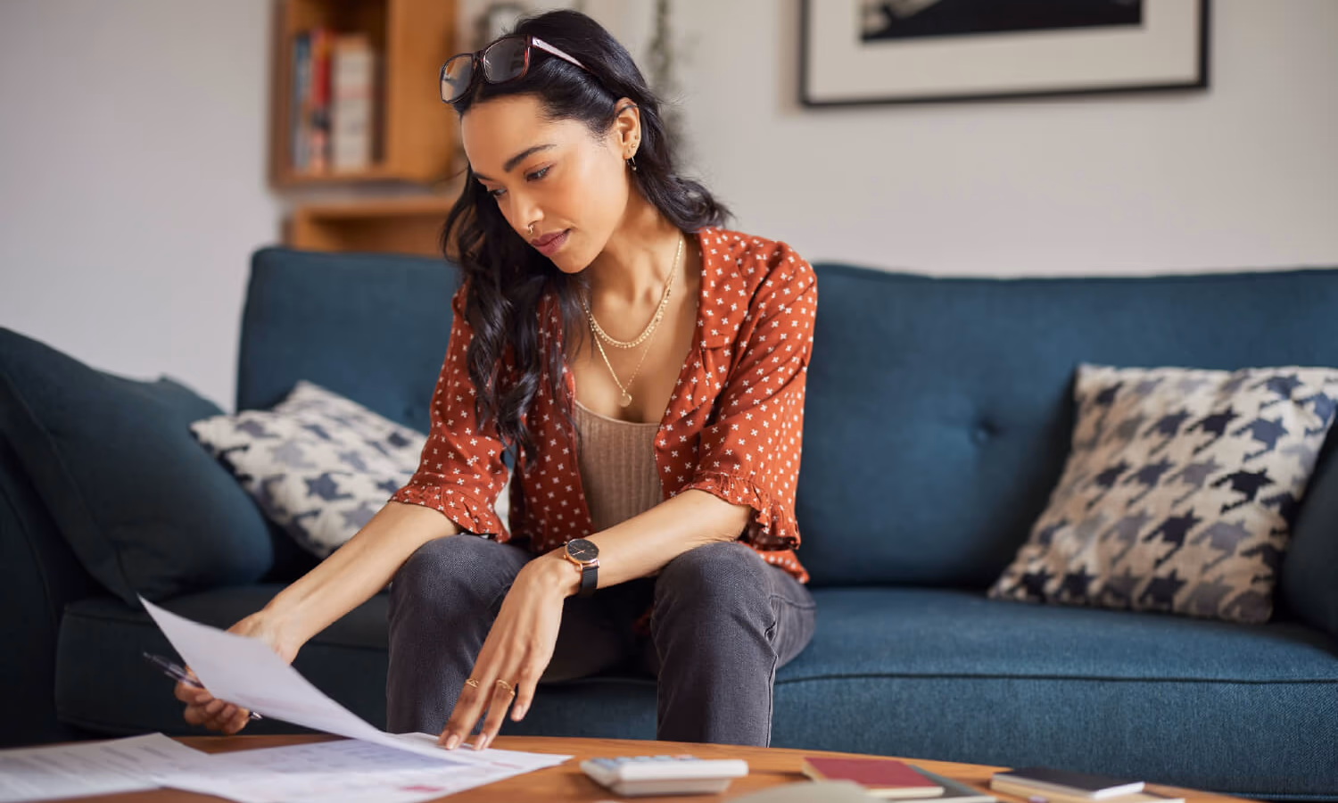 A woman sitting on a couch looking at documents laid out on a table with a calculator and notebooks.