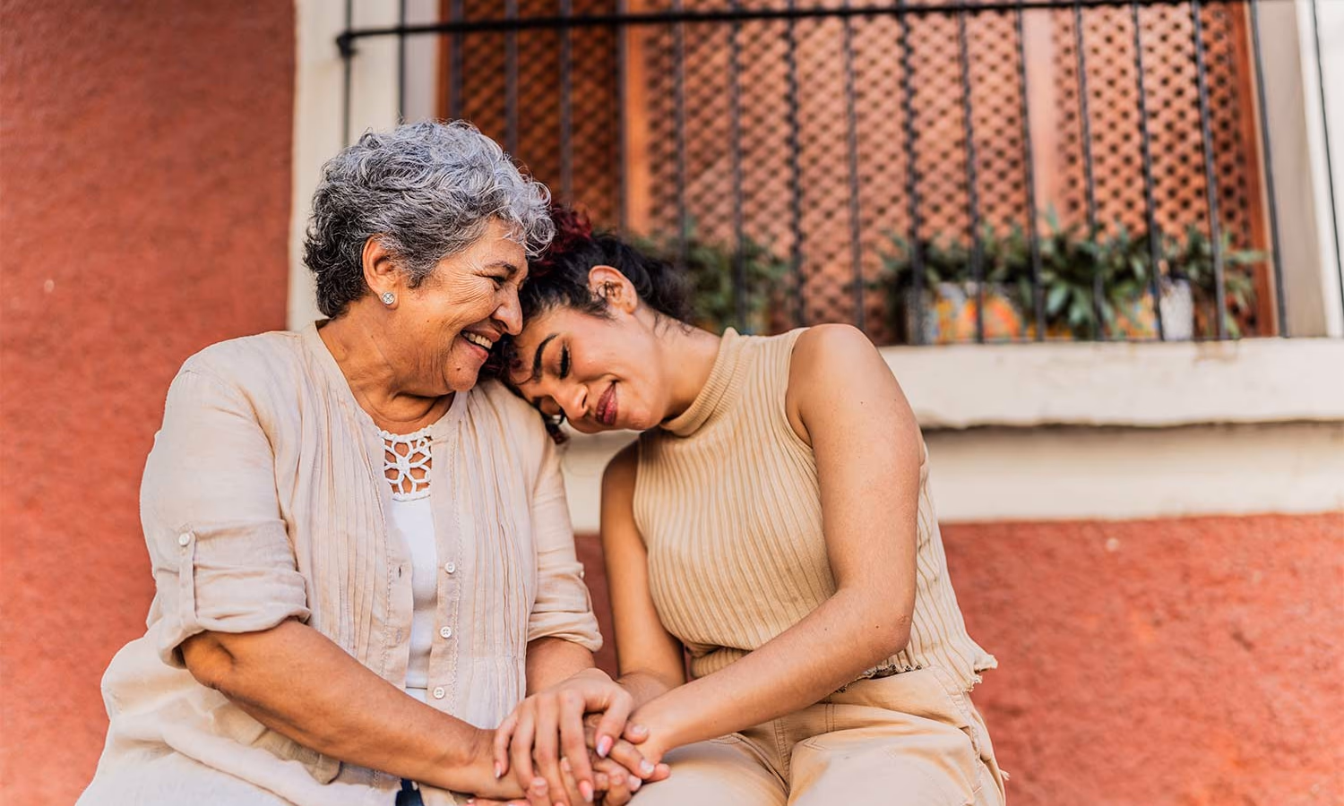 A mother and adult daughter sitting together, holding hands. They're smiling and the daughter's head is on the mother's shoulder