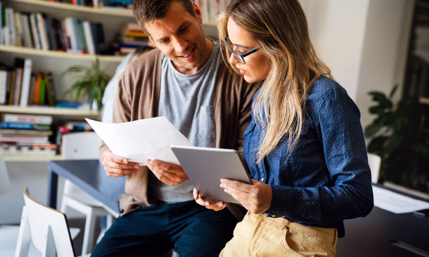Two people convene to review a sheet of paper and a tablet.