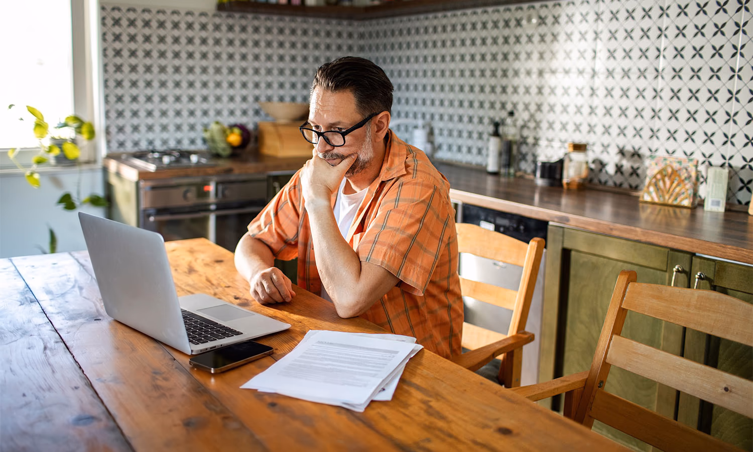 A man sits at a kitchen table, concentrating on the laptop and documents in front of him.