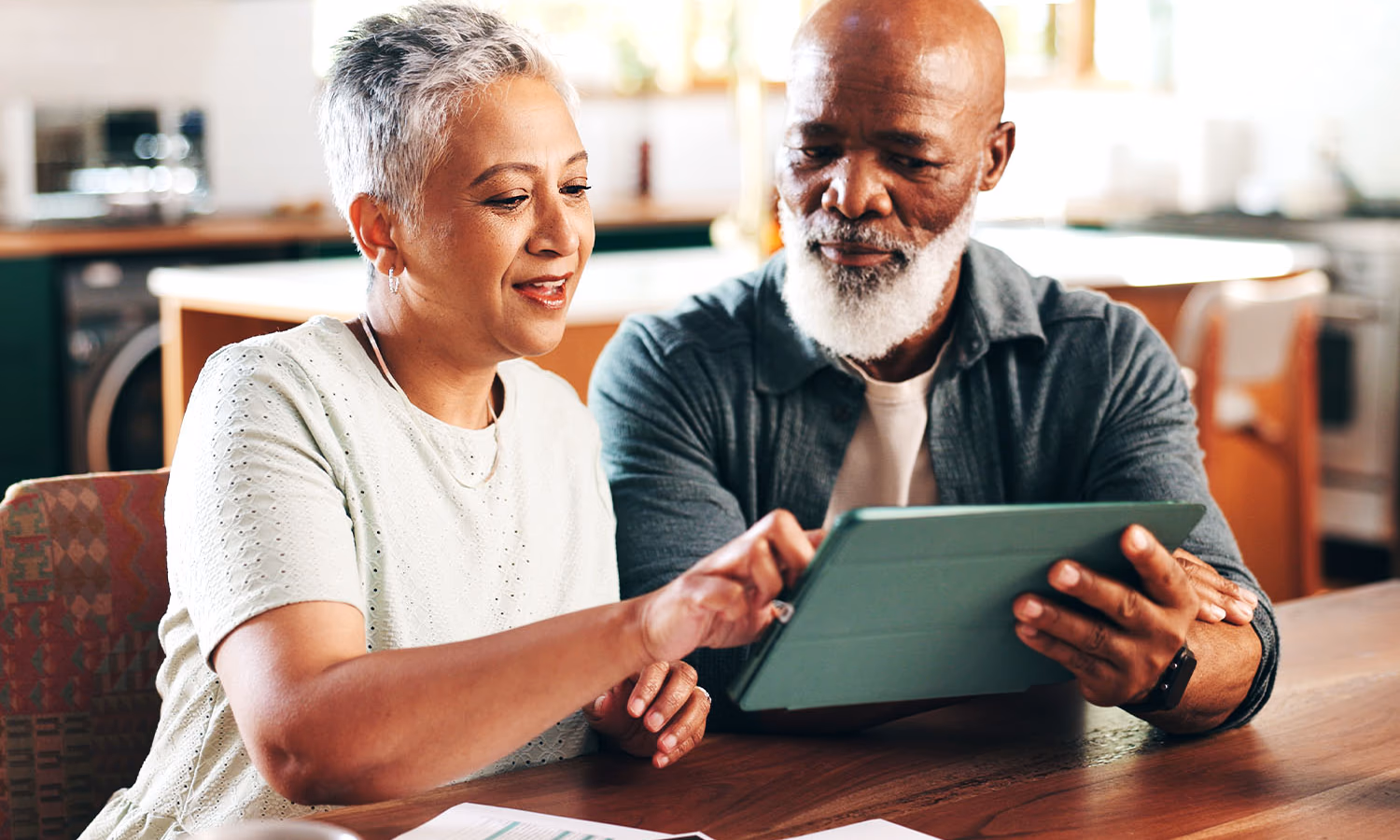 A couple sitting in their kitchen, leaning on one another and reviewing a document on a tablet.