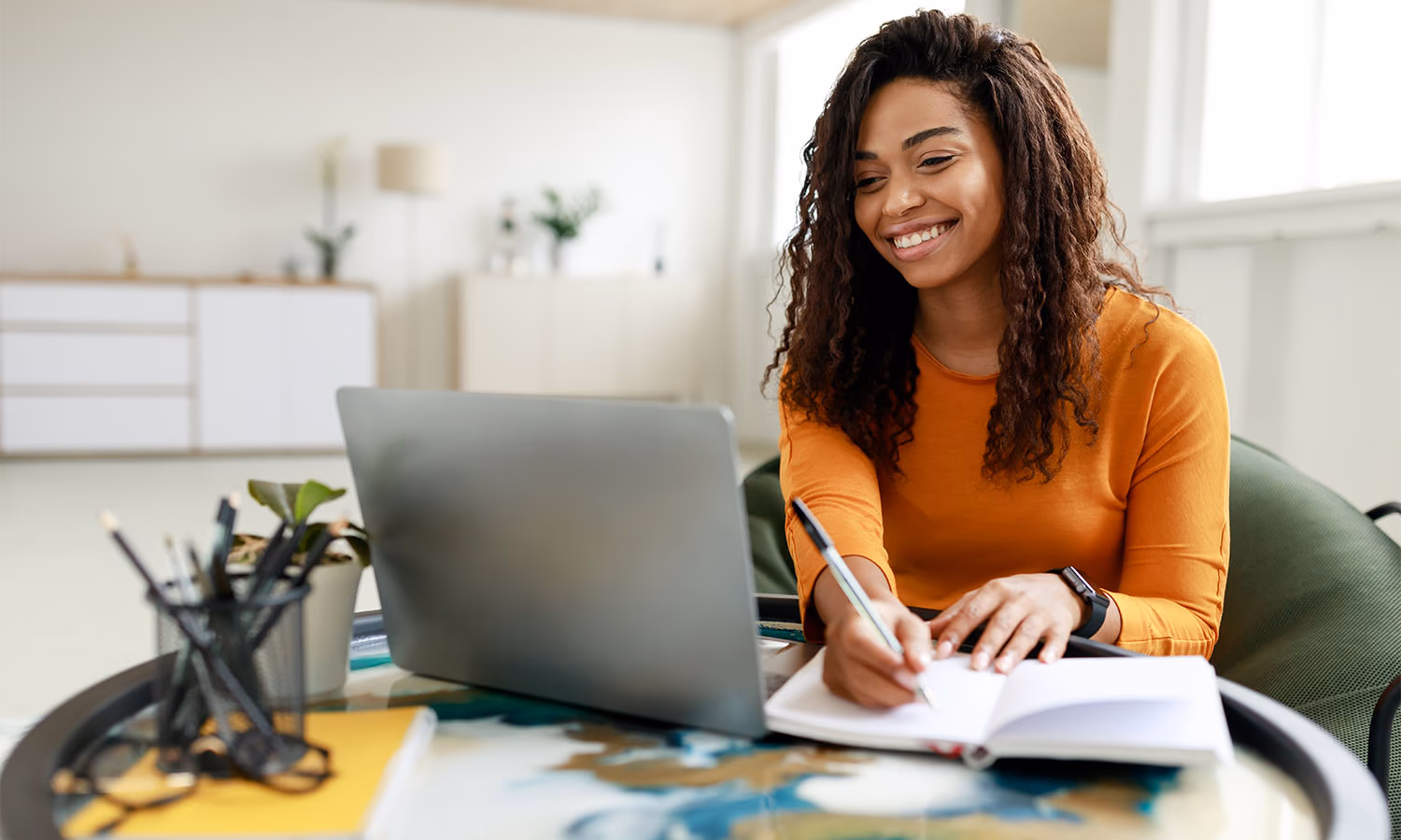 A woman sits at a table with a laptop in a room with bright daylight.