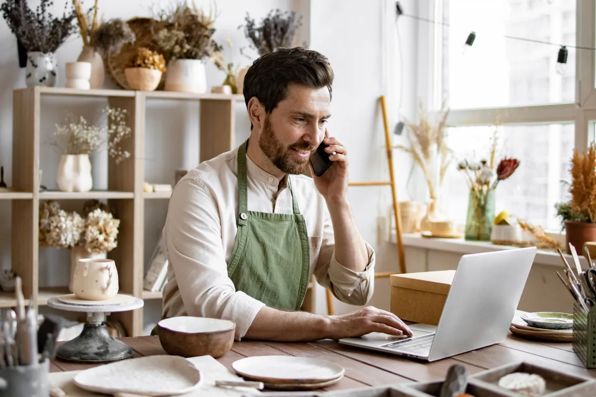 A man sitting in his pottery shop working on a laptop.