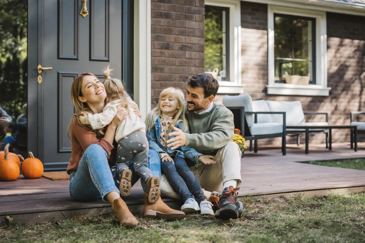 A family of two adults and two small children sits embracing on the front porch of a home.