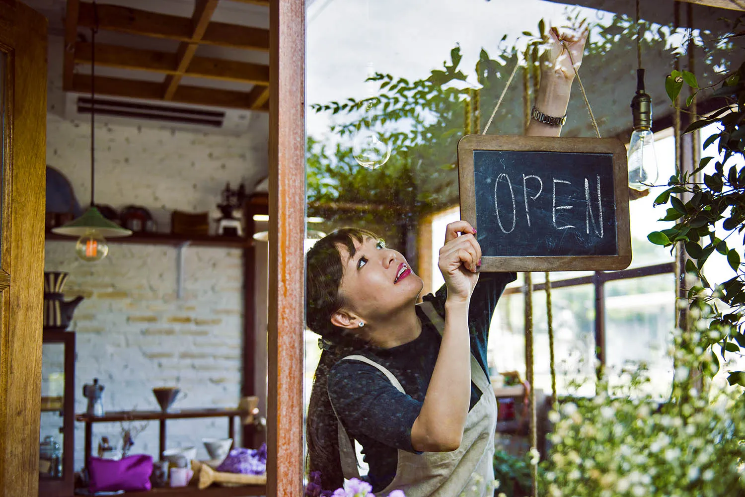 A person wearing an apron holds open the door to a cafe and flips around the "open" sign in the window.