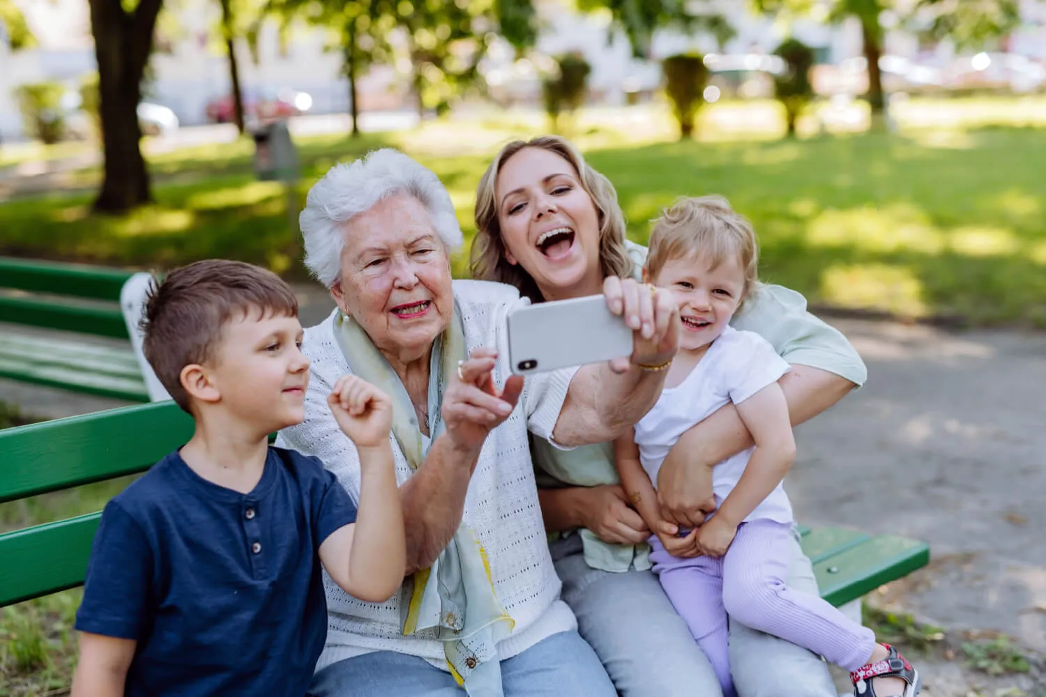 A multi-generational family sitting on a park bench on a sunny day, taking a photo together.