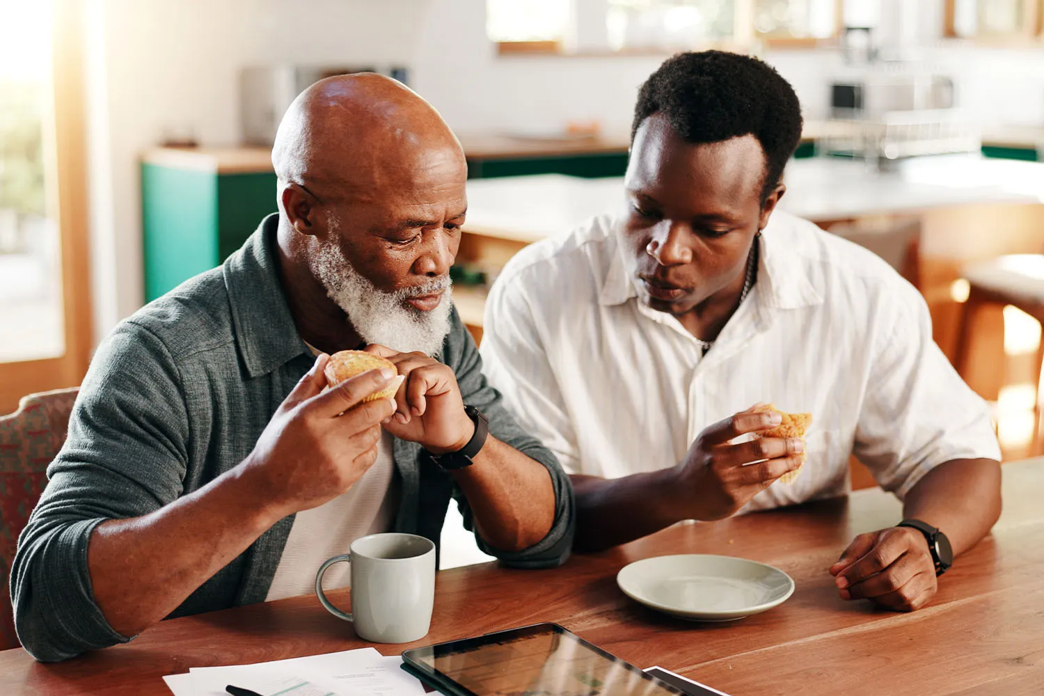An adult and a senior relative look at a document on a tablet while snacking on bread.
