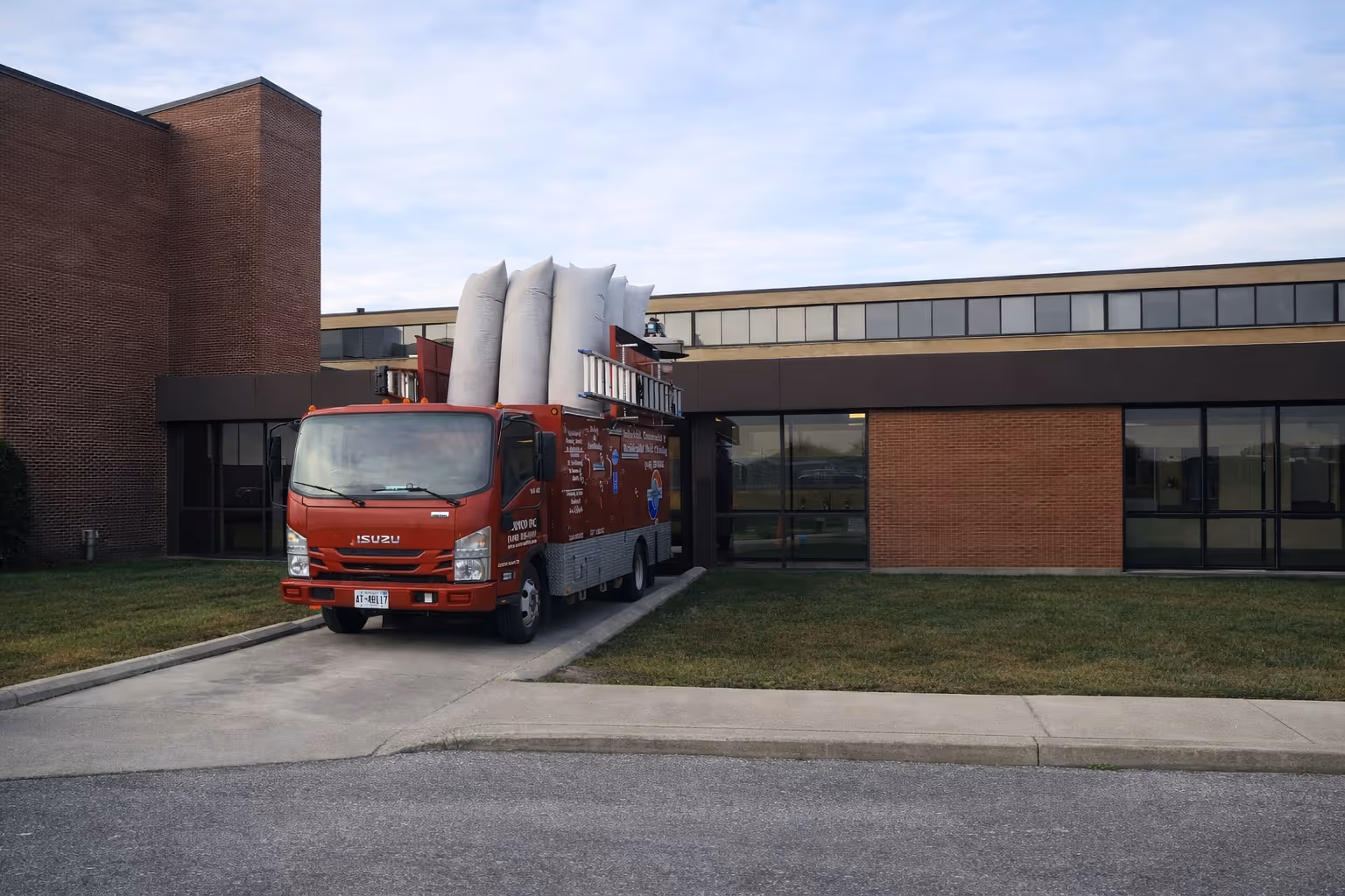 Red Isuzu truck parked on a driveway next to a brick building with large windows, carrying large white bags and ladders.