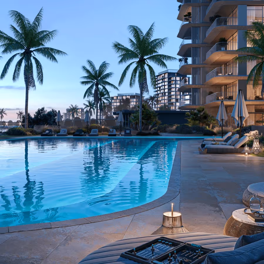 Luxurious outdoor swimming pool area at dusk with palm trees, modern apartment balconies, loungers, umbrellas, and a backgammon board on a table.