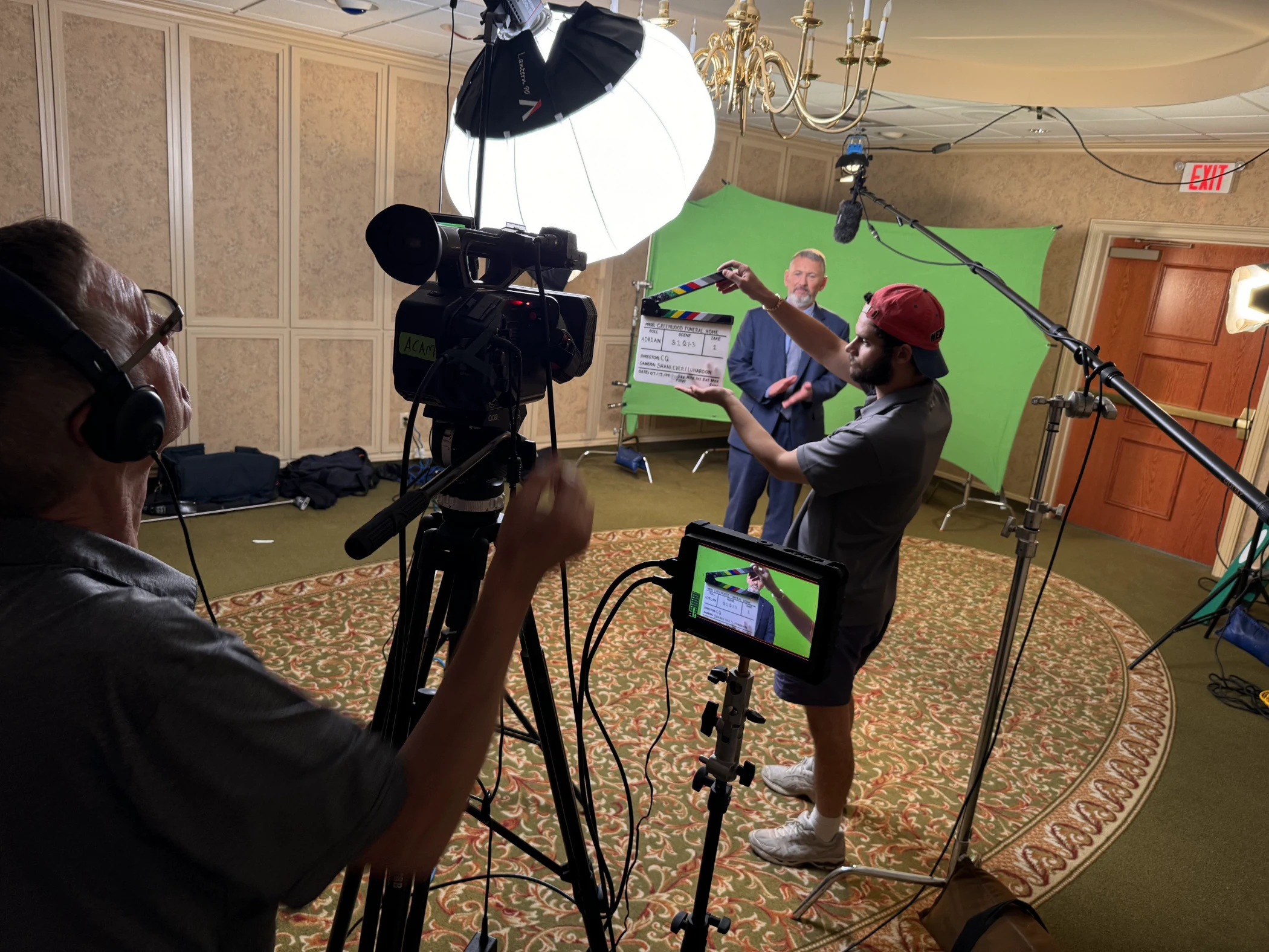 Man holding a clapperboard in front of a camera operator filming a man in a suit standing against a green screen backdrop in a room with professional lighting and audio equipment.