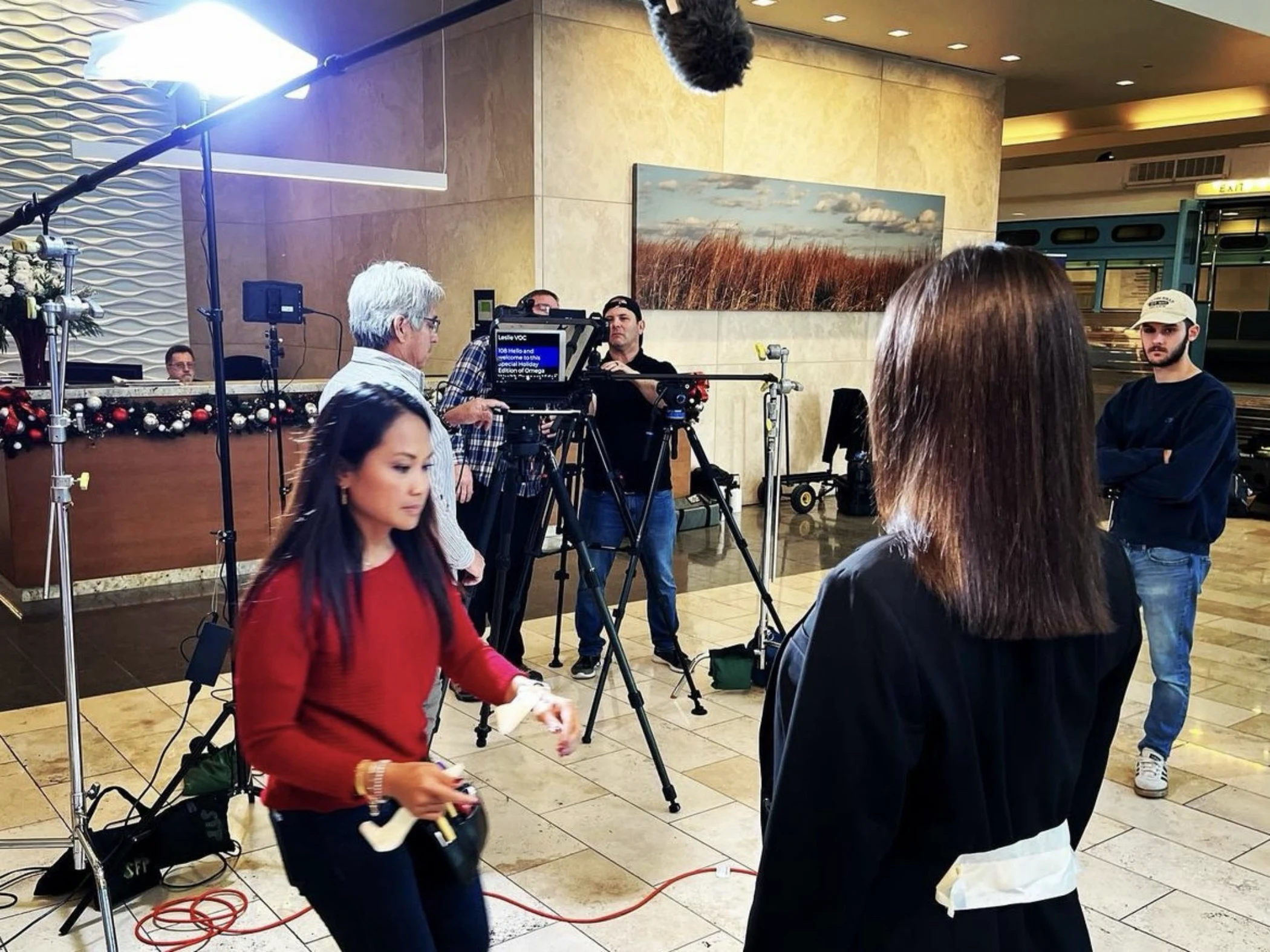 Film crew setting up cameras and lighting equipment in an indoor lobby with a woman in a red sweater walking by and a woman in a black jacket facing away.