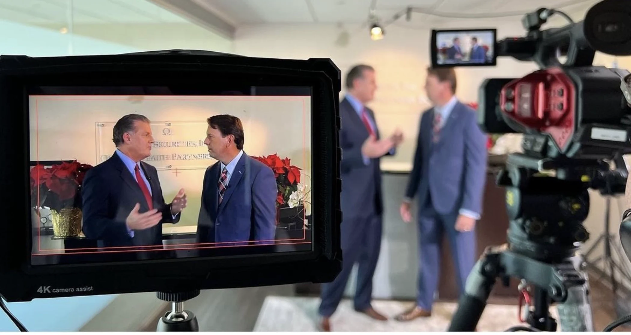 Camera screen showing two men in suits having a conversation indoors with poinsettia plants in the background.