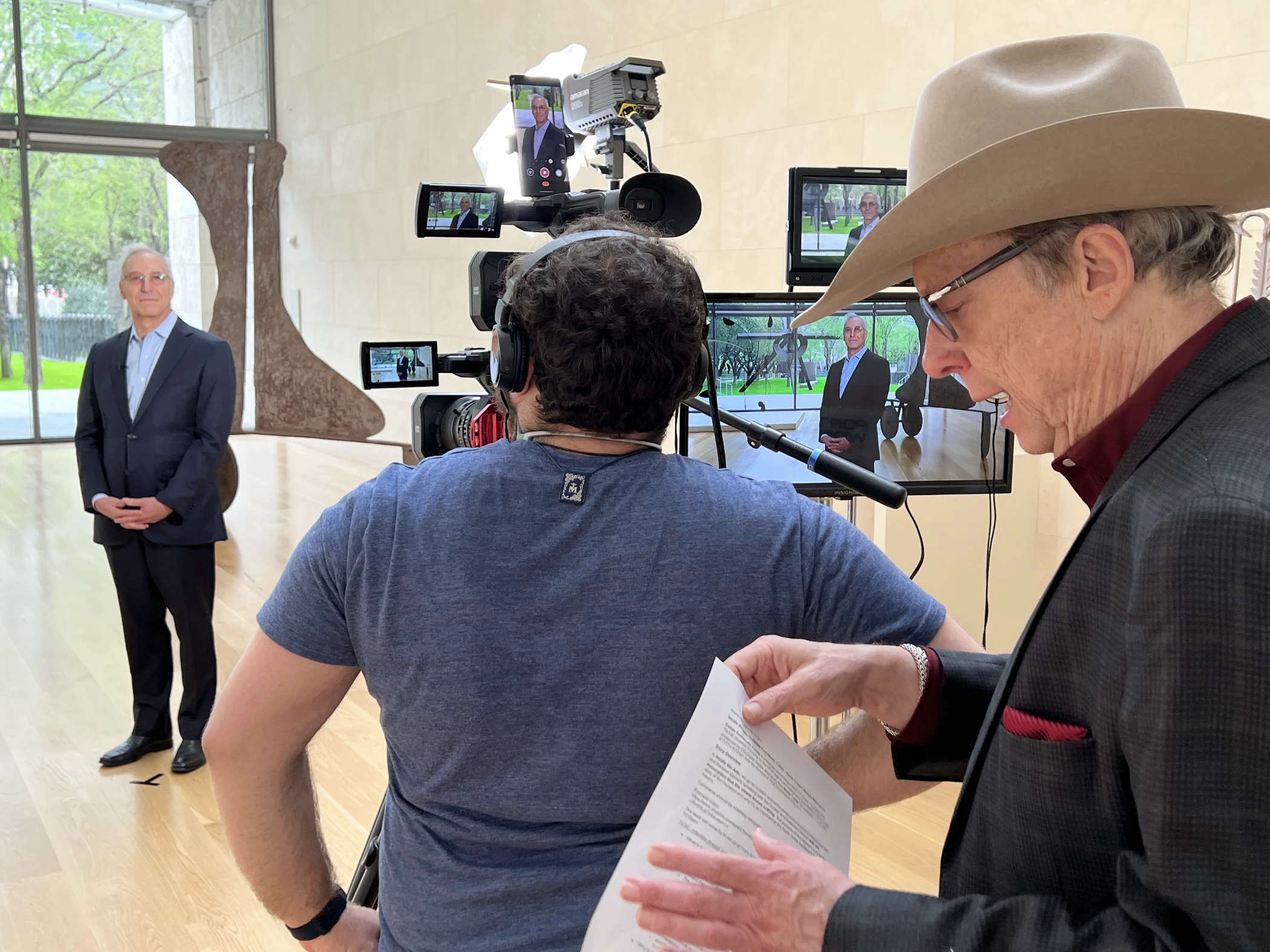 A cameraman filming a man in a suit standing in a gallery with a sculpture, while another man in a hat reads a script nearby.