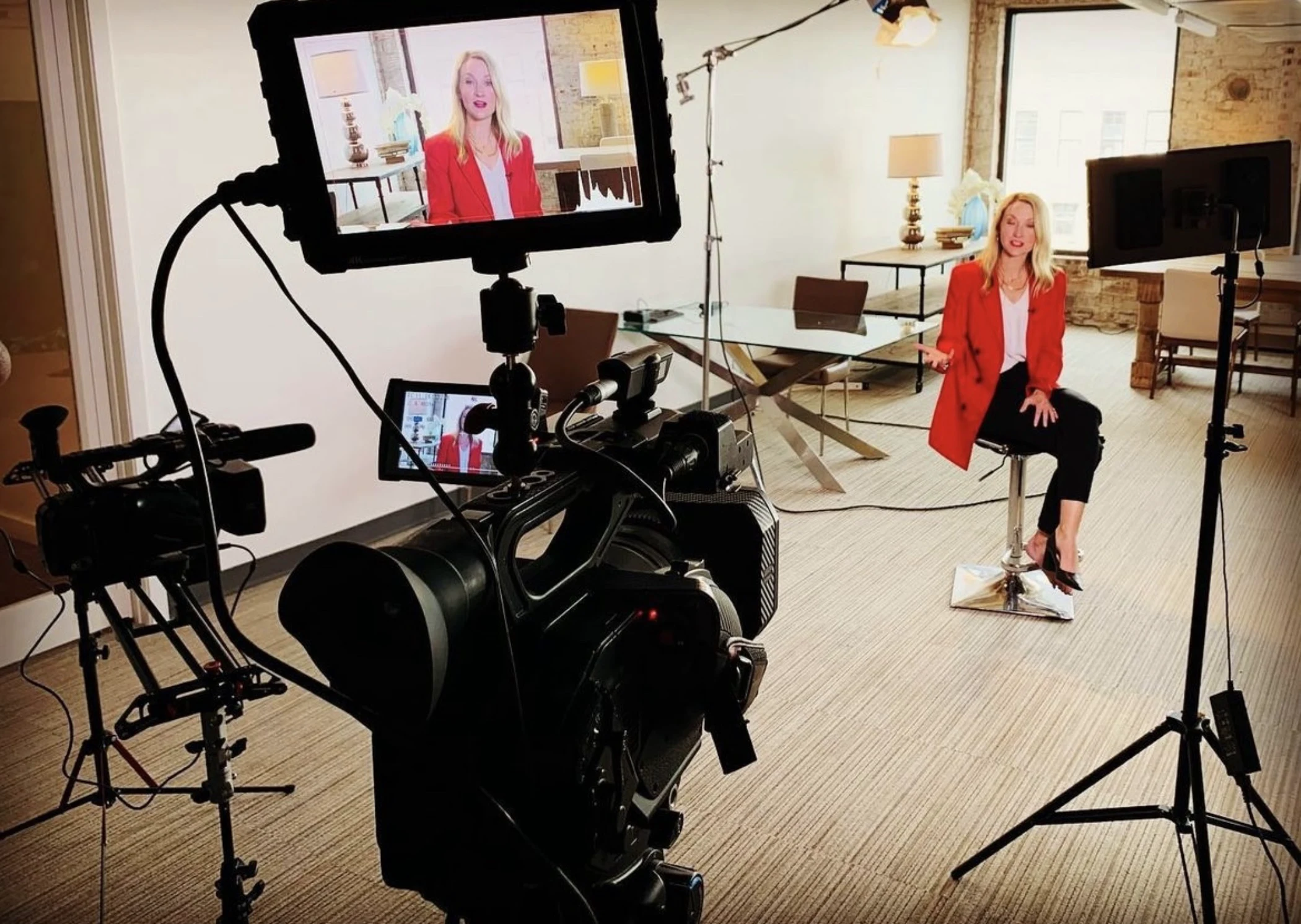 Woman in a red blazer sitting on a stool being filmed by professional video cameras and lights in a bright room.