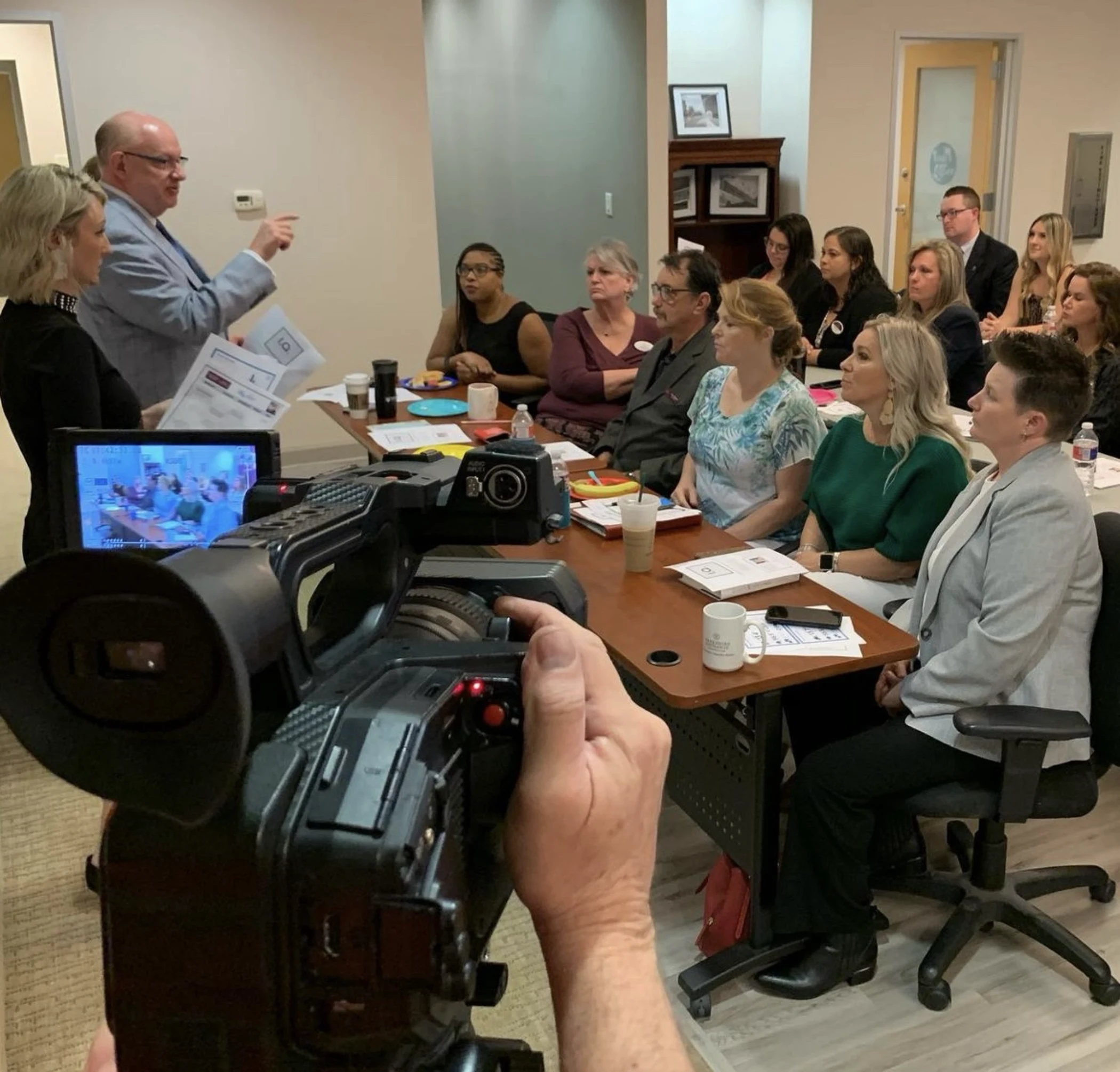 A group of people seated around a conference table attentively listening to a man in a light blue suit speaking, with a video camera filming the scene.