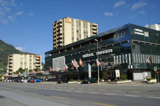 Modernes mehrstöckiges Gebäude mit Glasfassade und der Aufschrift 'Möbel Furrer' sowie ein hoher Hotel- und Appartementturm im Hintergrund.