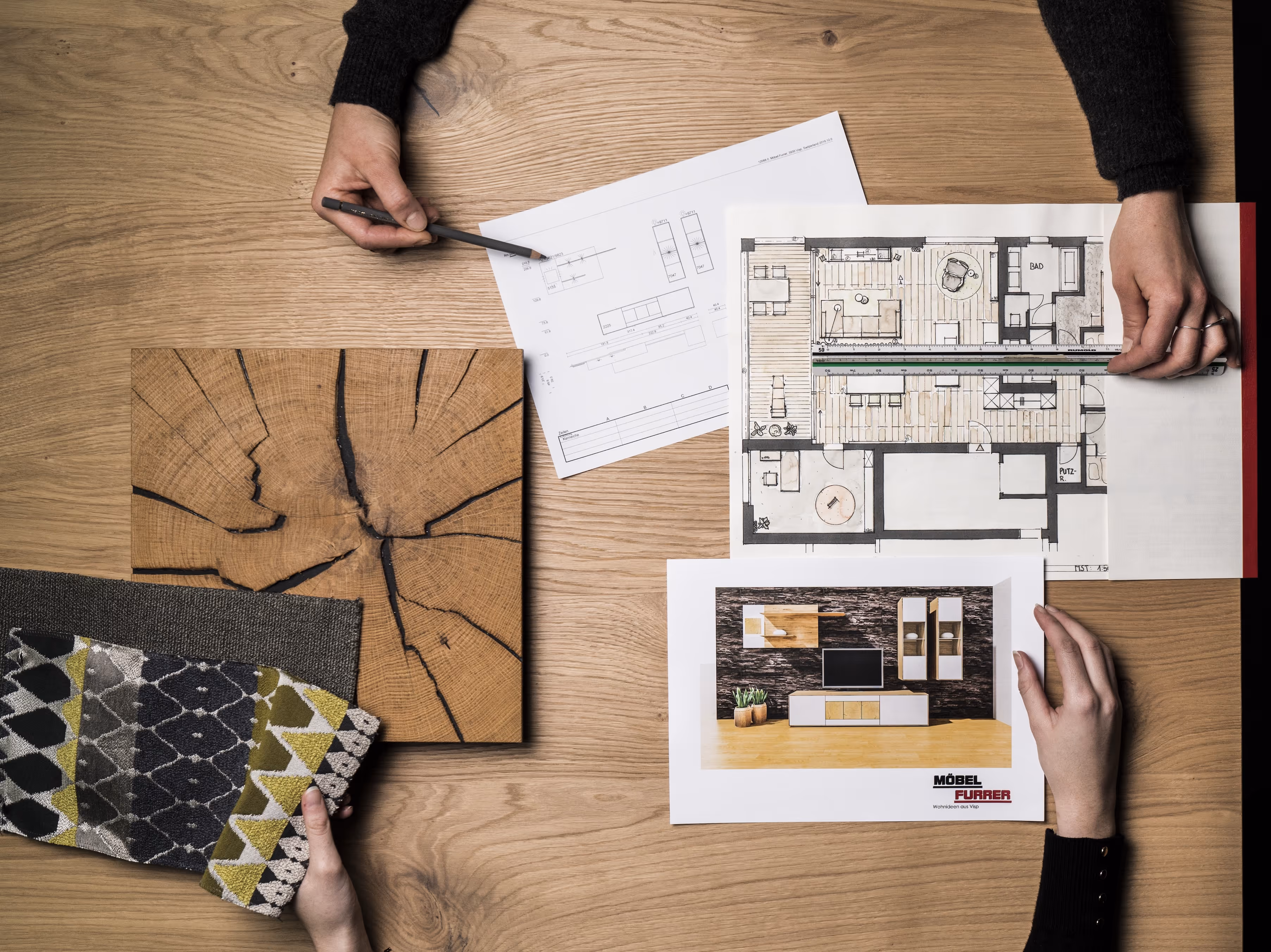 Drei Hände arbeiten an einem Holztisch mit Bauplänen, einem Musterstück Holz und verschiedene Stoffmustern für Innenarchitektur.