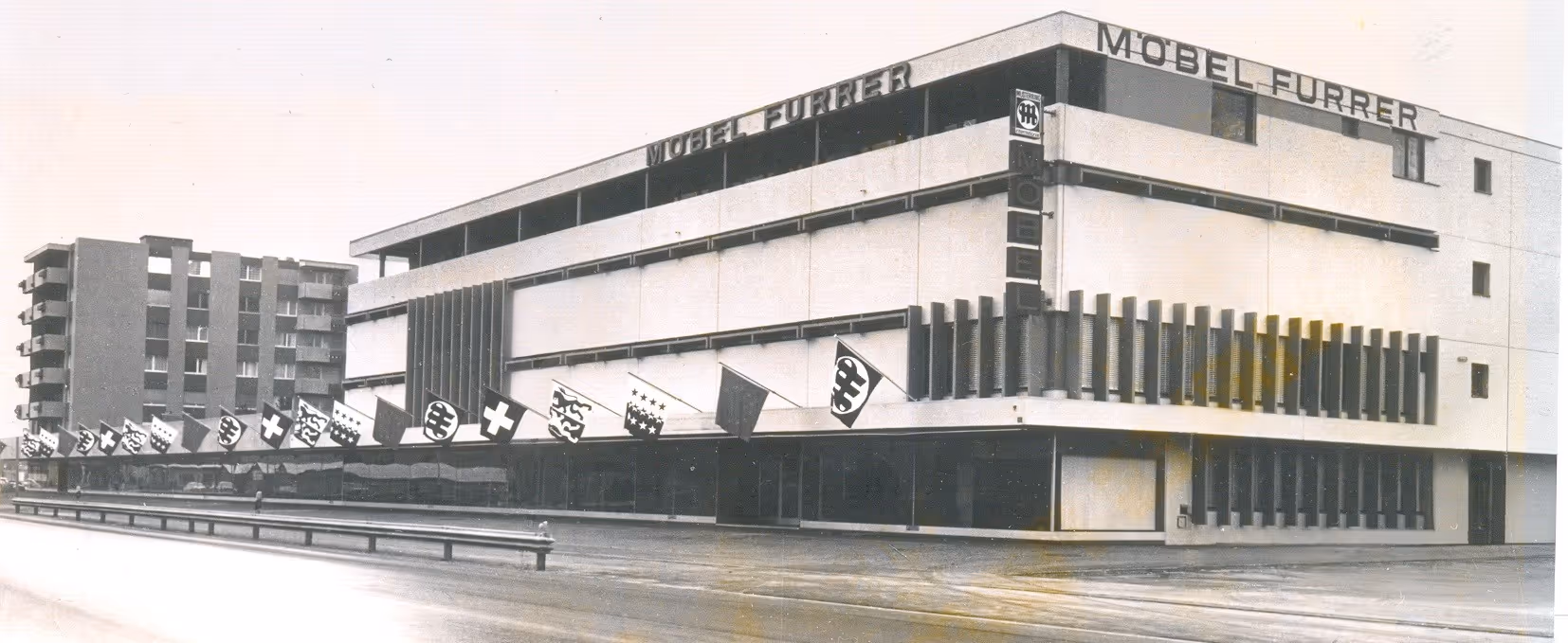 Historisches Schwarz-Weiss-Foto eines grossen modernen Gebäudes mit der Aufschrift 'Möbel Furrer' und mehreren Fahnen entlang der Fassade.