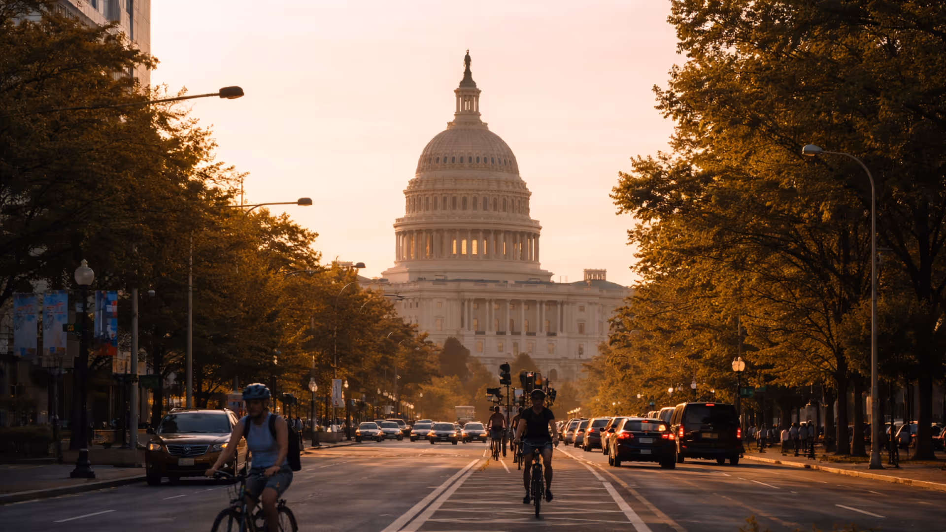 The United States Capitol building at sunset with cyclists and cars on the street lined with trees.