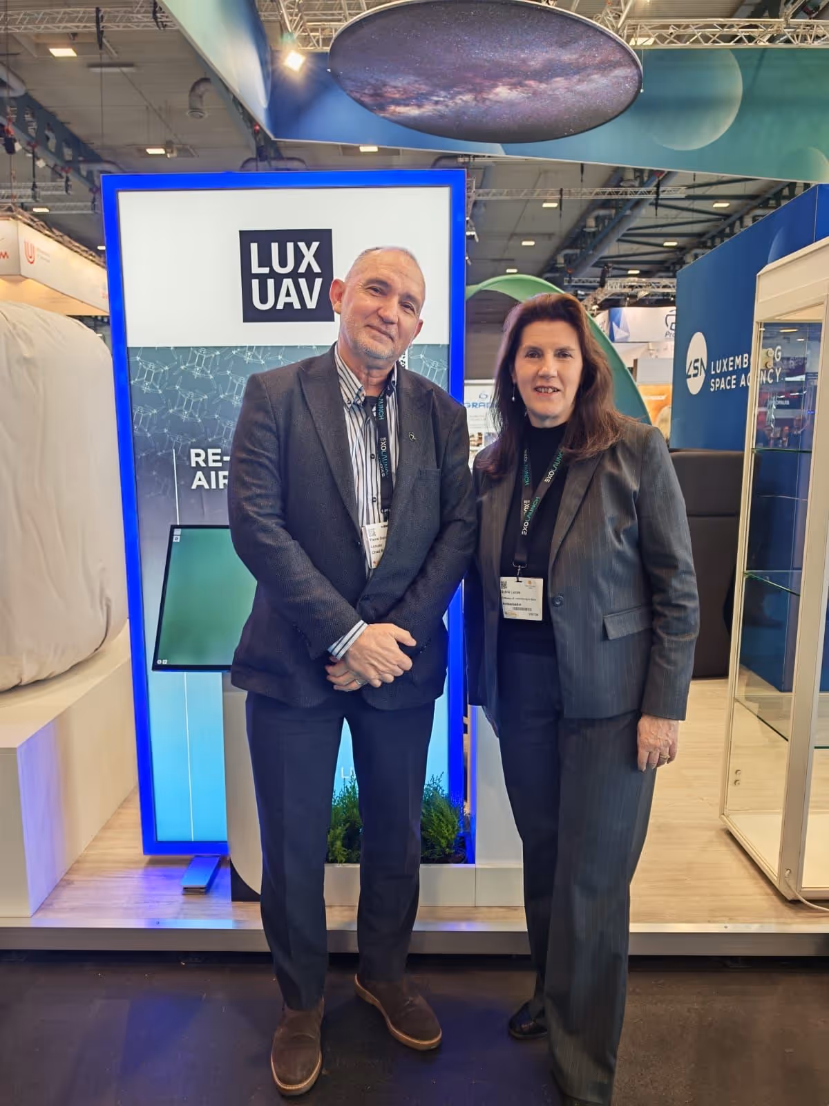 Man and woman in business attire standing in front of a LUXUAV display at a trade show.