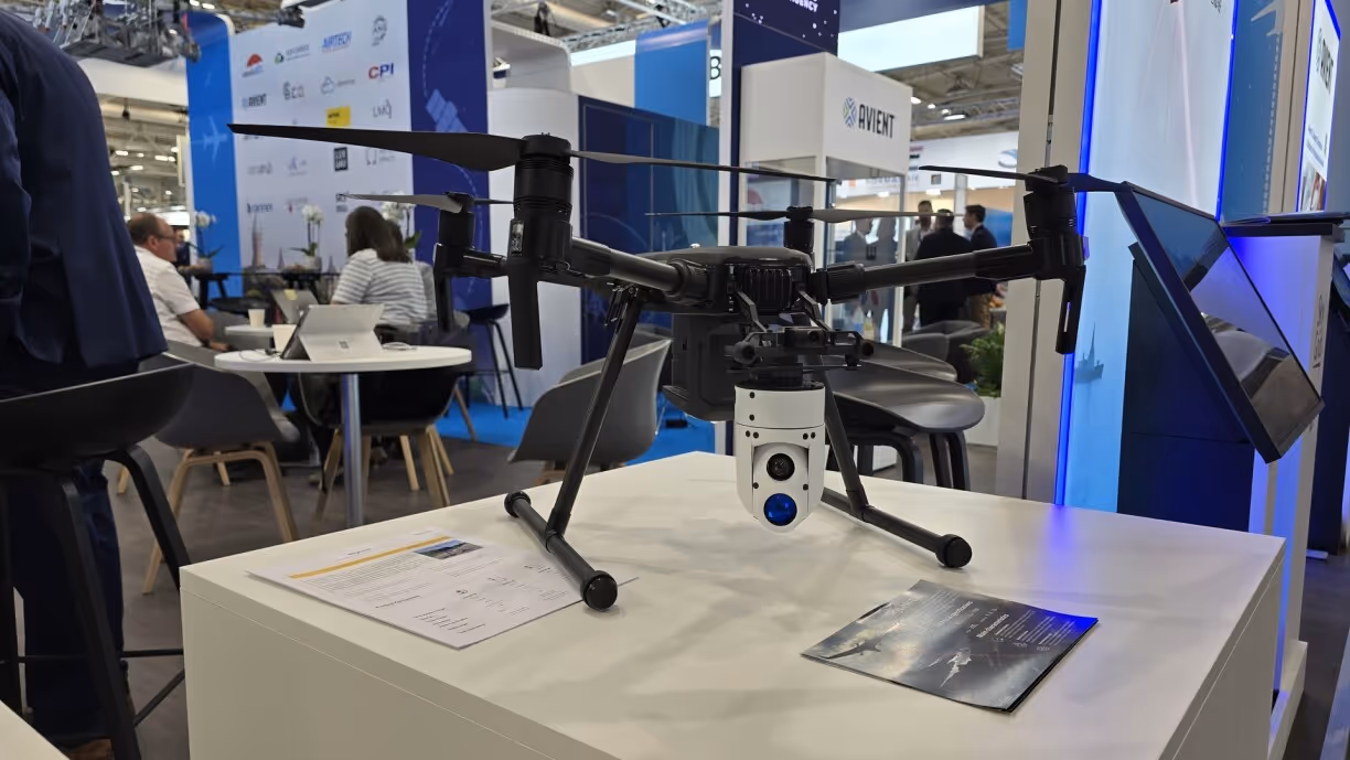 Black quadcopter drone with a camera mounted on a white display table at a technology exhibition.