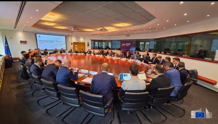 Large group of people in business attire seated around an oval conference table in a modern meeting room with a European Union flag and logo visible.