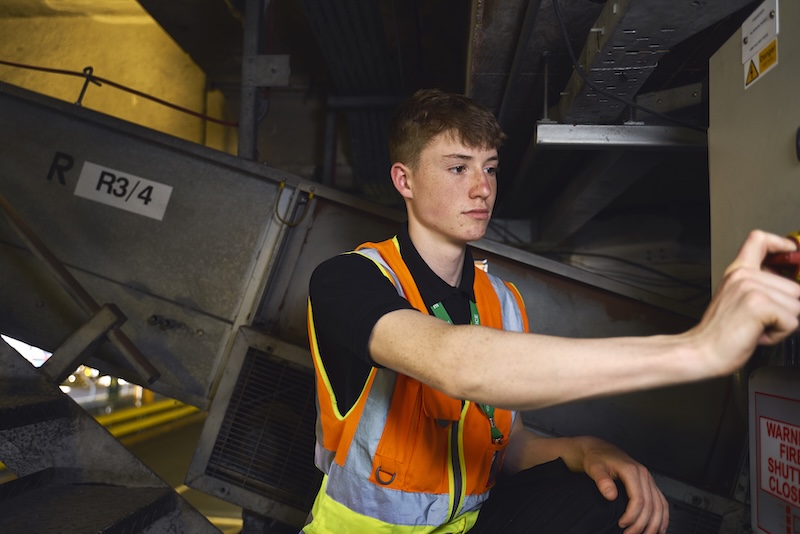 Young male worker in an orange safety vest operating industrial machinery indoors.